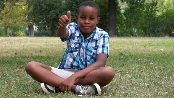 A Young  Black Boy Sits on Grass in a Park and Shows a Thumb Up To the Camera with a Smile alt