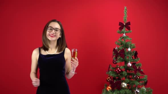 Young Woman Dancing with a Glass of Champagne Near the Christmas Tree on a Red Background. A Girl alt