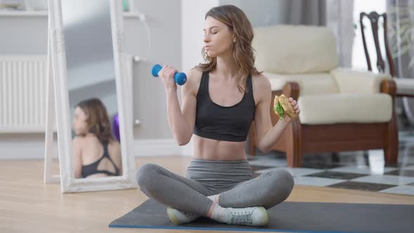 Front View of Slim Woman Eating Unhealthy Food Lifting Dumbbell Sitting on Exercise Mat alt