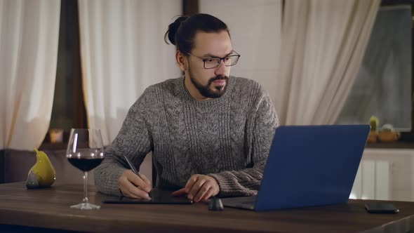 A Young Handsome Bearded Man Works on a Laptop with a Glass of Wine