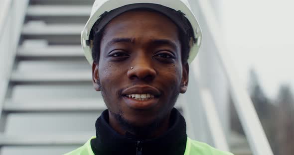 A Man in Work Uniform Smiles Looking at the Camera Standing Near a Wind Turbine alt