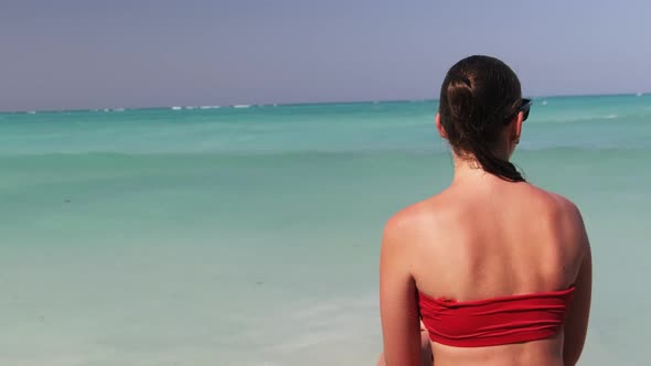 Young Woman in Sunglasses and Red Bikini Sitting and Looking at Ocean Rear View alt