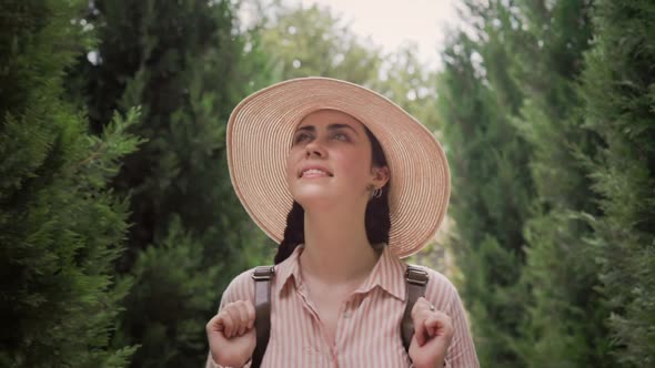 Happy beautiful young woman in a straw hat is walking down the street, among the trees. Follow shot. alt