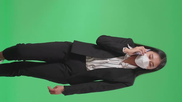 Asian Business Woman Wearing Mask Talking On Mobile Phone While Walking In Front Of Green Screen alt