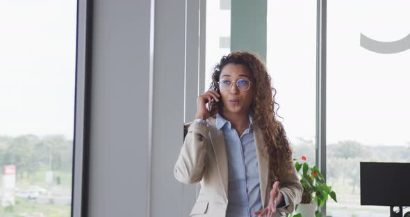 Biracial businesswoman sitting by desk and talking on smartphone in modern interiors alt