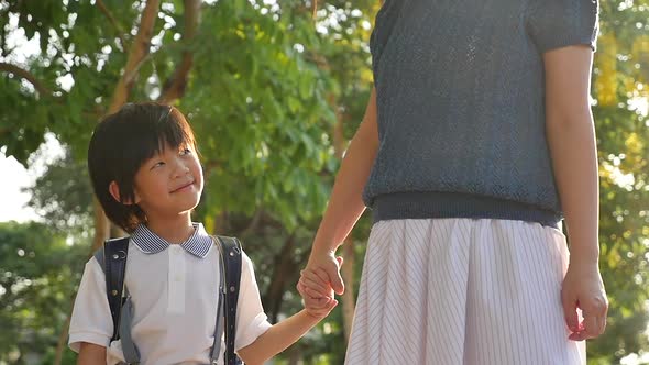 Asian mother holding hand of little son with backpack outdoors, back to school slow motion alt