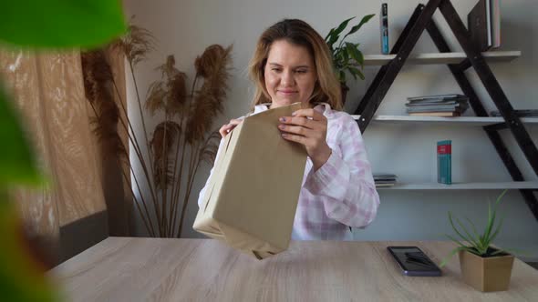 A Young Caucasian Lady is Sitting at the Table at Home and Opens a Box alt
