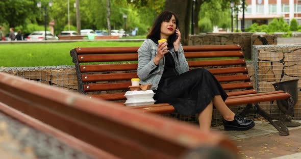 Woman sitting on the bench in the park, talking on mobile phone, with take away food and coffee alt