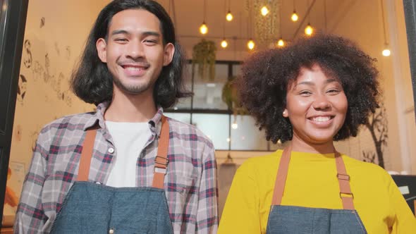 Two baristas at a cafe door, arms crossed, look at a camera with welcome smile. alt