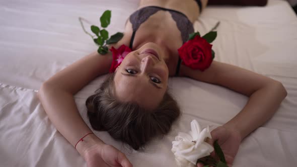 Young Passionate Happy Woman Looking at Camera Lying on Bed with Flowers Around alt