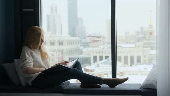 caucasian woman reading a travel book sitting on the windowsill