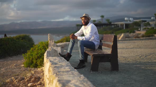 Side View of Thoughtful African American Gay Man Sitting in Sunset Rays on Bench Looking Away alt
