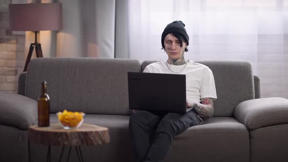 Confident Concentrated Tattooed Man Sitting on Couch Surfing Internet on Laptop alt
