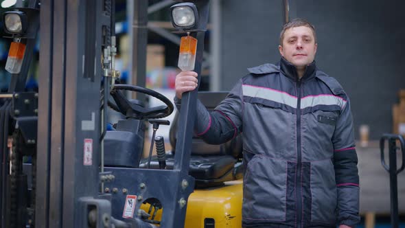 Confident Male Driver in Uniform Posing at Forklift in Warehouse Indoors alt