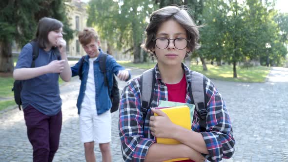 Genius Schoolboy in Eyeglasses Standing on Schoolyard As Blurred Classmates Laughing and Pointing at alt