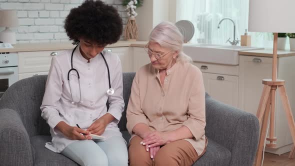 Afro American Woman Doctor Writes Prescription for Senior Woman Sitting on Couch at Home alt