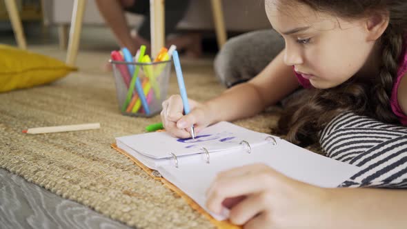 Teenager Girl Draws in Notebook with Pen Lying on Floor