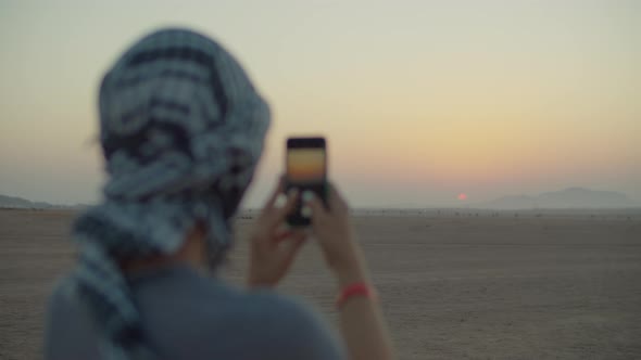 Woman in Checkered Keffiyeh Taking Picture of Sunrise on Mobile Phone Standing in the Desert alt