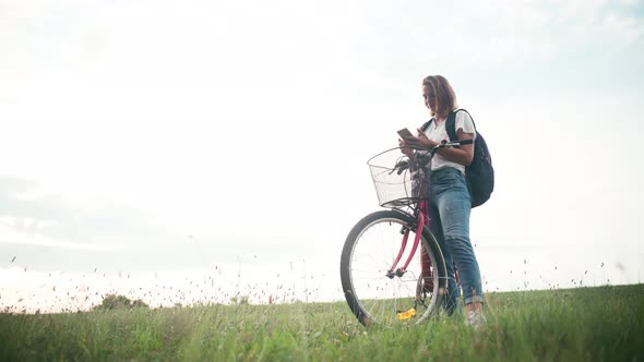 A Young Cheerful Woman with a Backpack Standing in the Field with Her Bicycle alt