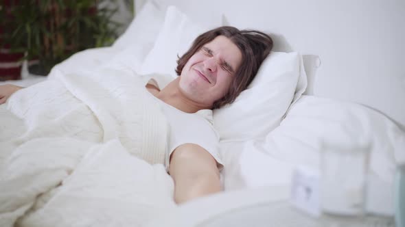 Sleeping Young Man Looking at Clock Smiling and Stretching Lying in Bed in the Morning alt