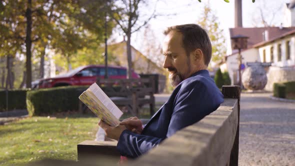 A Middleaged Handsome Caucasian Man Reads a Book with a Smile As He Sits on a Bench