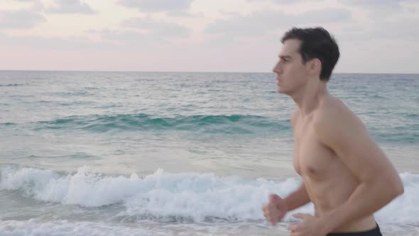 Track shot on shirtless male runner jogging on beach in the evening alt