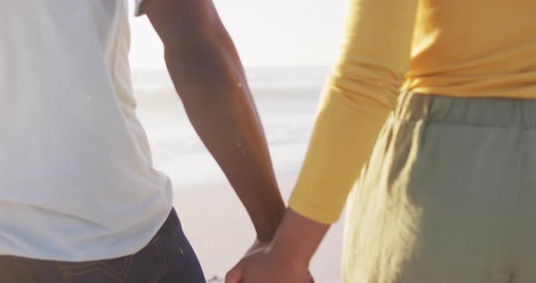 Smiling african american couple walking and holding hands on sunny beach alt