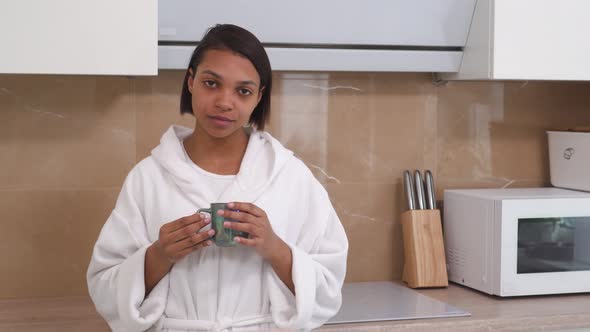 Young Woman Drinking Coffee in the Morning in a White Coat in the Kitchen