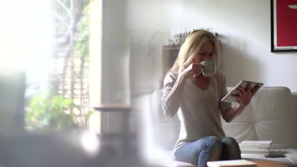 Mature woman drinking coffee and using digital tablet in living room