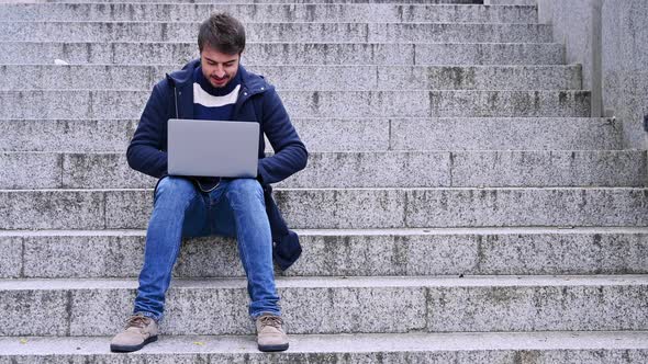 Young Bearded Businessman Sits on Steps, Using Laptop and Looks on His Screen. Hipster Man Is alt