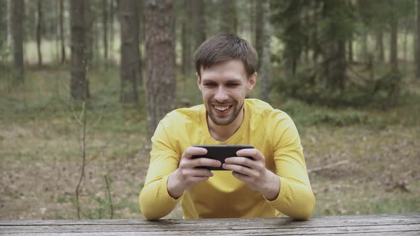 Satisfied Man in Yellow Longsleeve Plays Mobile Game on Smartphone in Forest alt