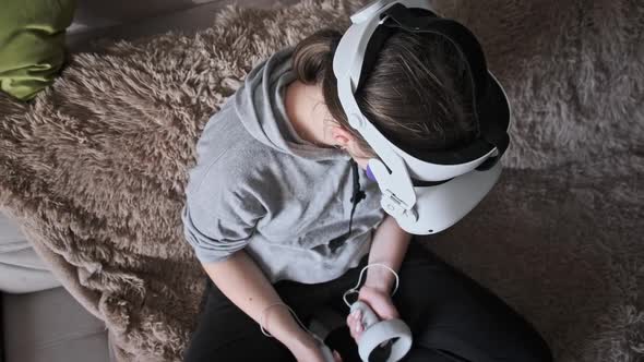 Young Woman Sitting on Sofa Playing Game Using VR Helmet Indoor at Home alt