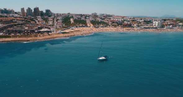 Drone Flies Around Sailing Yacht with Beach and Cityscape View on Summer Day