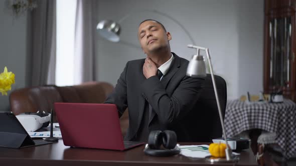African American Young Man Stretching Neck Sitting at Table in Home Office Indoors alt