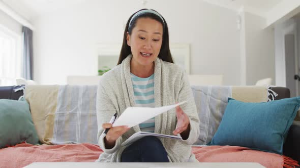 Asian woman working at home, sitting on couch making video call, talking and reading paperwork alt