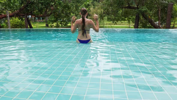 A Woman Sits on the Edge of the Pool and Swims Against the Backdrop of Tropical Vegetation in a