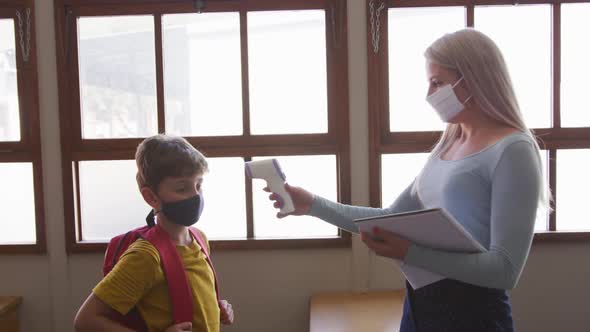 Female teacher wearing a face mask measuring temperature of a boy in class at school alt
