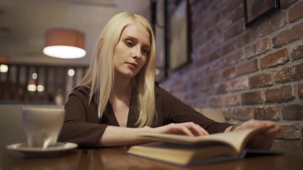 A Young Blonde Woman Enthusiastically Reads an Interesting Book in a Coffee Shop