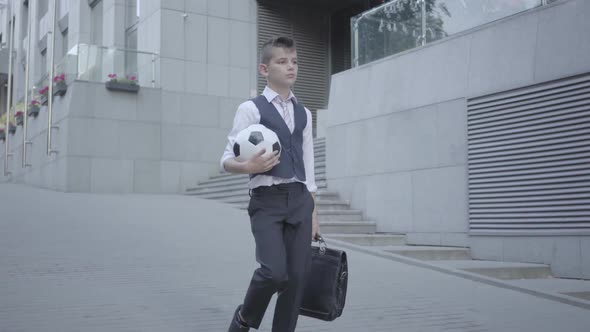 Serious Well-dressed Boy Walking Down the Street Holding the Soccer Ball and Purse in Hands alt