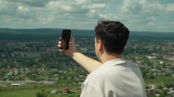 A Young Adult Male Takes a Smartphone Picture From the Top of a Large Hill alt