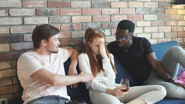 Redhead Woman Crying Sitting on the Bed with a Glass of Champagne Among Two Different Men. alt