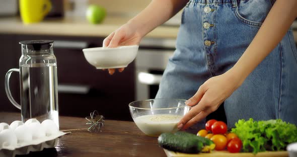 Young Woman is Stirring Eggs and Oatmeal with a Whisk in a Transparent Bowl