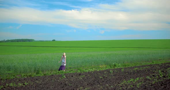 A Girl in a Dress and a Hat Walks Through a Green Field of Grass.