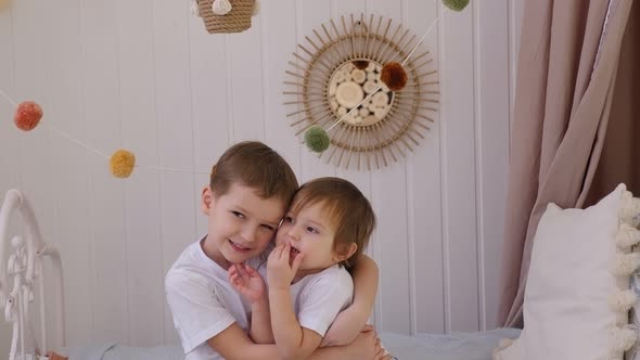 Brother and Sister Cuddle Sitting at Home on the Bed in the Bright Bedroom alt