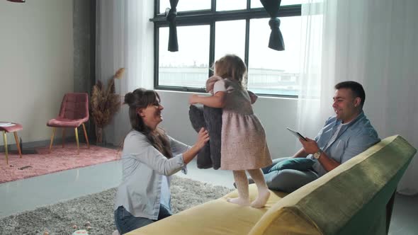 Little Girl Playing with Mother with Pillows While Her Father Works Using a Tablet alt