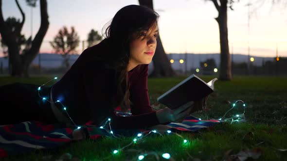 A cute young woman reading a fictional story book outdoors at twilight with lights glowing SLIDE RIG alt