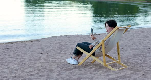 Woman video chatting saying hello on phone, sitting on a beach alt