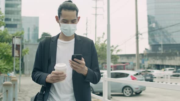 Asian businessman wearing a surgical mask and using smartphone while walking on the street.