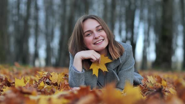 A Young Girl in an Autumn Park Lies on the Leaves and Smiles with a Leaf in Her Hand. alt