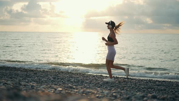 A Young Woman Runner is Listening to Music in Earphones and Training By a Sea alt